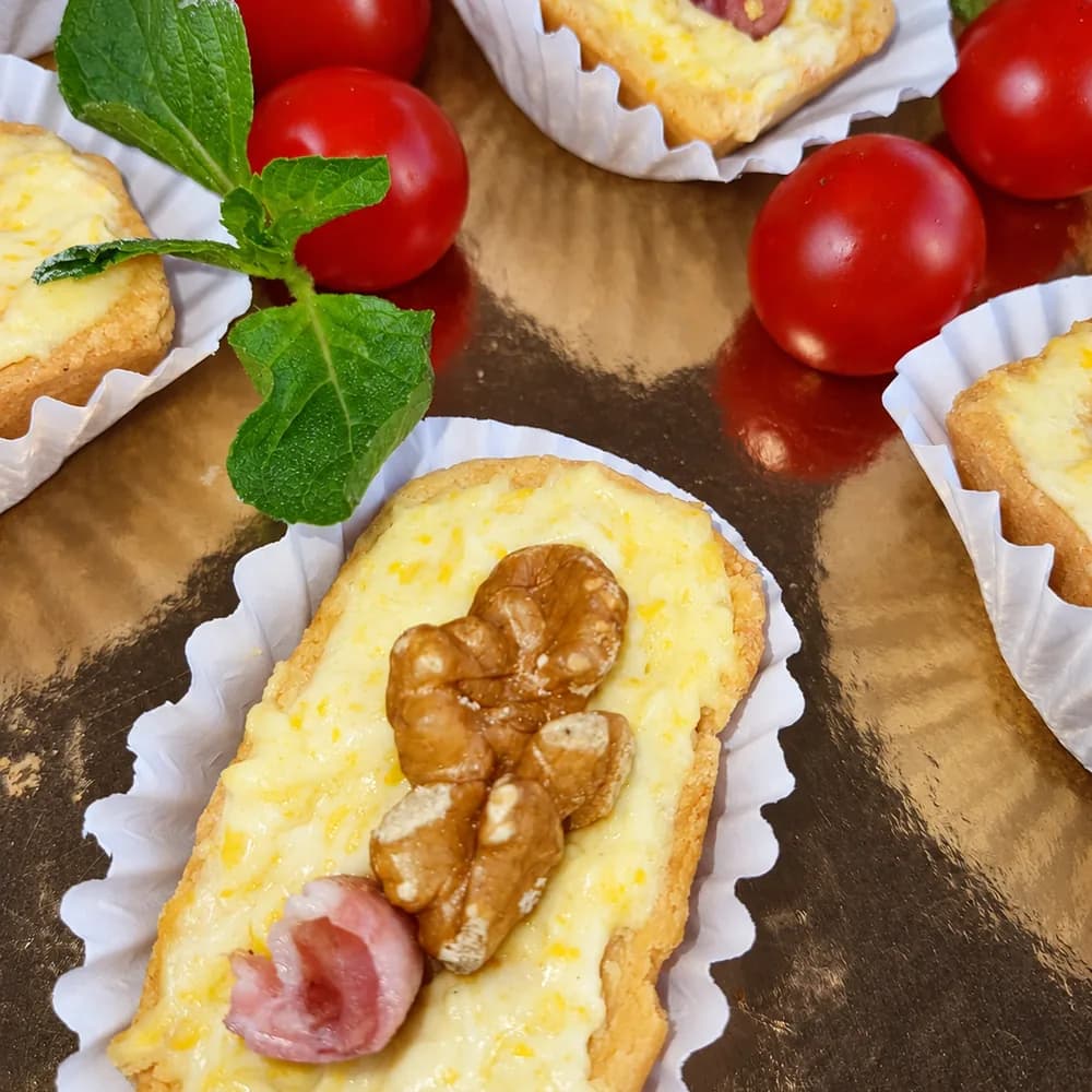 Bocaditos salados en pirotines de papel, cubiertos con queso y decorados con una nuez y jamón, acompañados de tomates cherry y hojas de albahaca